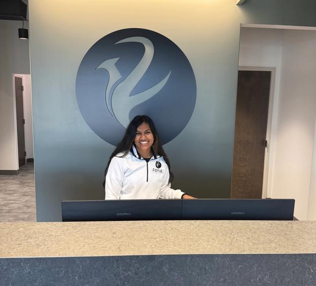 A smiling woman with long dark hair stands behind a reception desk, wearing a white shirt with a "RISE" logo. Behind her is a wall with a large circular logo.