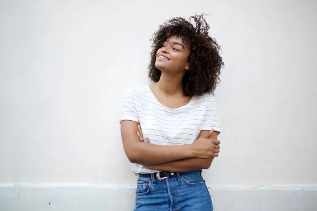 a woman with curly hair is standing in front of a white wall with her arms crossed and smiling .