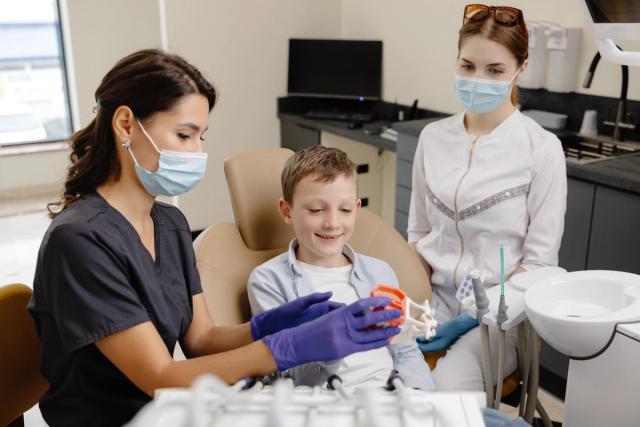 A masked dental professional shows a teeth model to a smiling boy in a dental chair, with another masked staff member in the background.