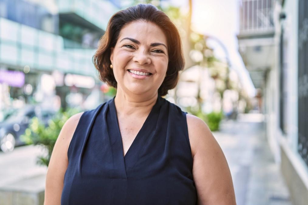 A smiling middle-aged woman with dark hair stands on a sunny city street.