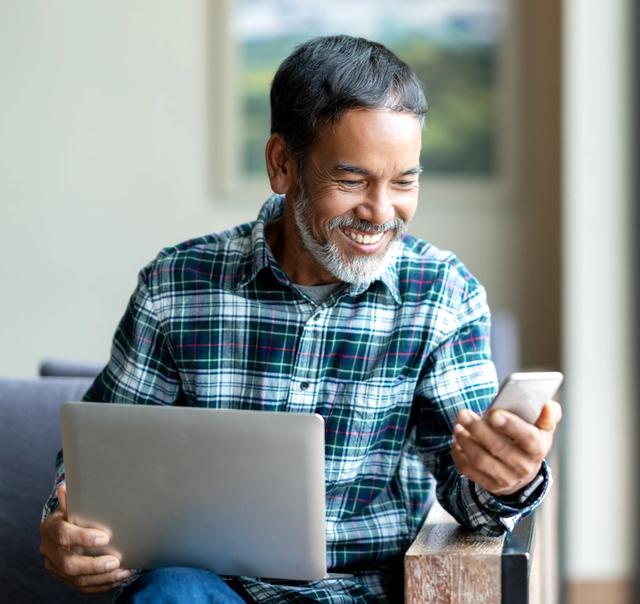 a man is sitting in a chair using a laptop and a cell phone .