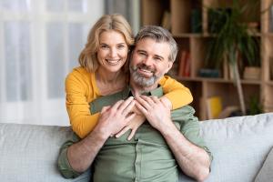 A smiling blonde woman hugs a bearded man from behind on a couch.