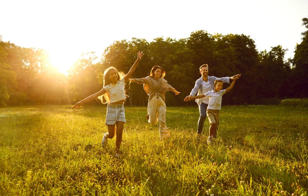 A family of four runs joyfully through a sunlit field with outstretched arms.