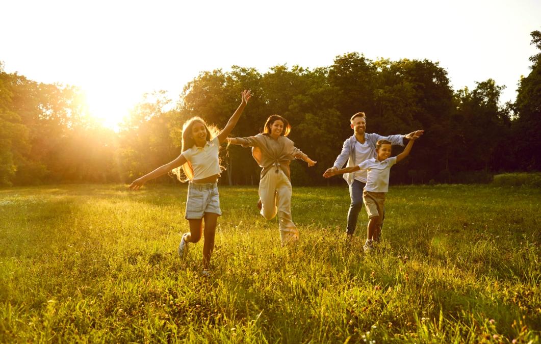 A family of four runs joyfully through a sunlit field with outstretched arms.
