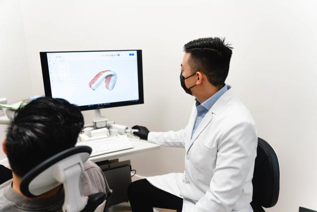 A masked dentist shows a patient a 3D dental model on a computer screen.