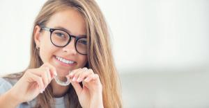 A smiling young woman in glasses holds a clear invisalign dental aligner.