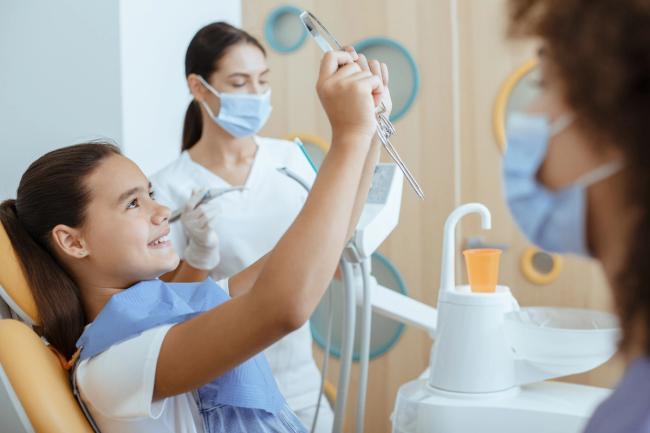 A smiling girl in a dental chair holds a mirror, with a masked dental professional standing behind her.