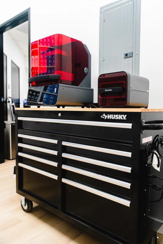 Two 3D printers, one with a red cover, on a black Husky tool chest.