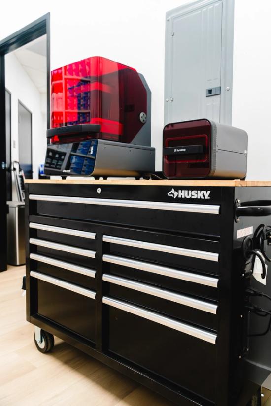 Two 3D printers, one with a red cover, on a black Husky tool chest.
