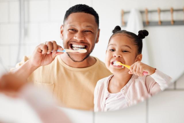 A father and daughter smile while brushing their teeth in a mirror.