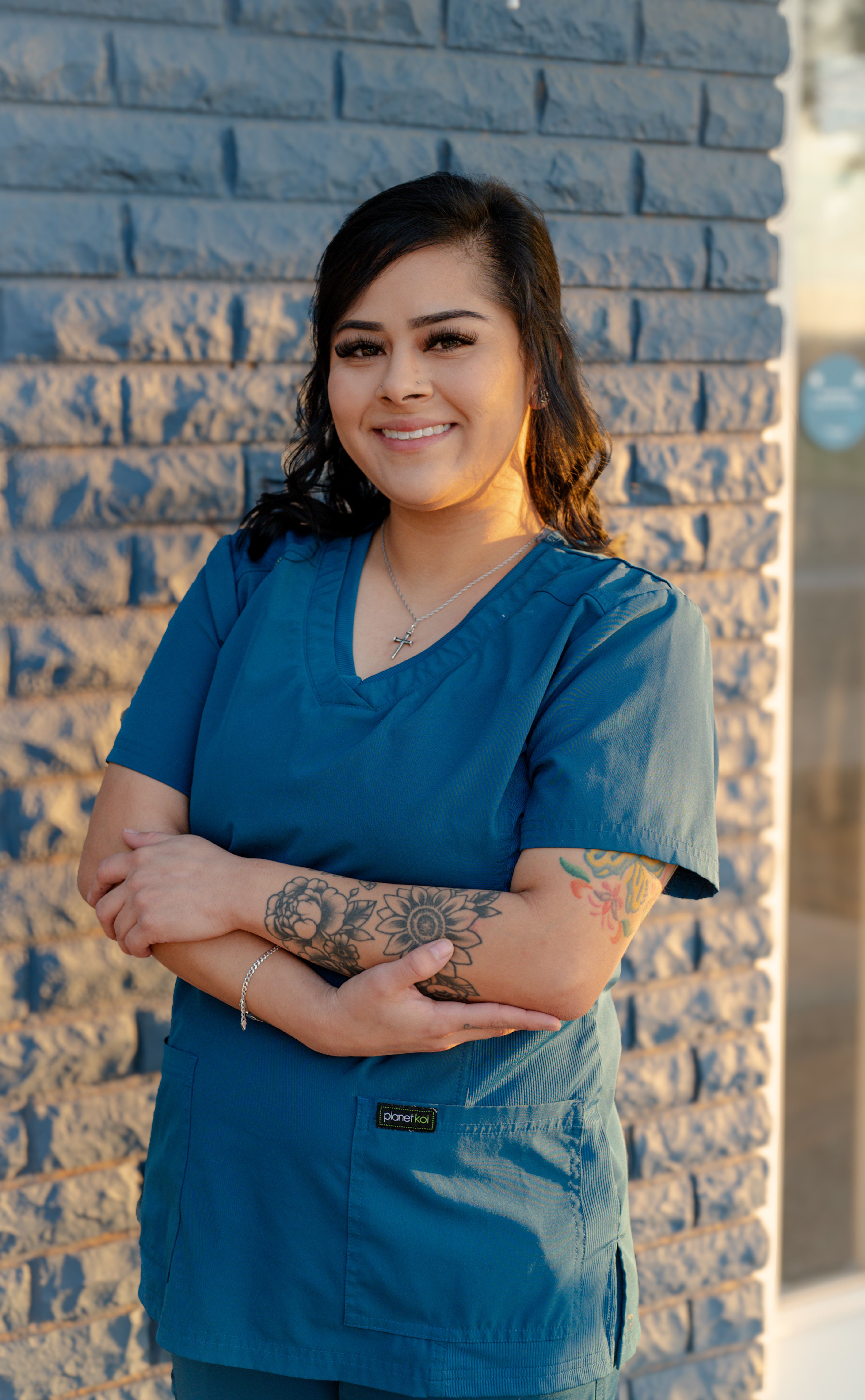 a woman in a blue scrub top with a tattoo on her arm is standing in front of a brick wall .