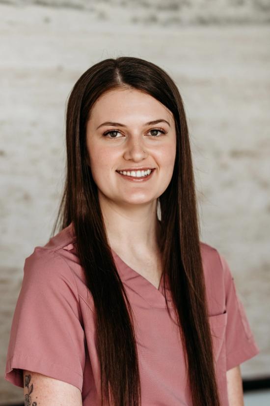 A smiling young woman with long dark hair, wearing a pink scrub top and a nose piercing.