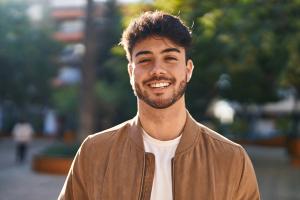 A smiling young man with dark hair and a beard wears a brown jacket outdoors.
