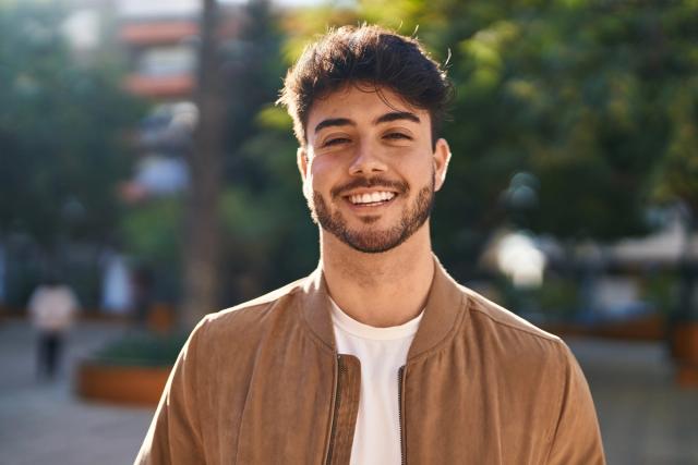 a young man with a beard is smiling for the camera while wearing a brown jacket .