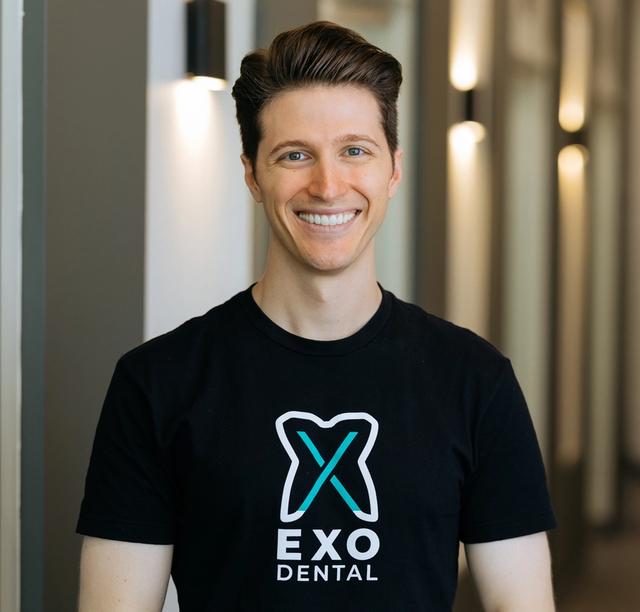 a man wearing a black exo dental t-shirt is smiling in a hallway .
