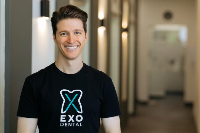 a man wearing a black exo dental t-shirt is smiling in a hallway .