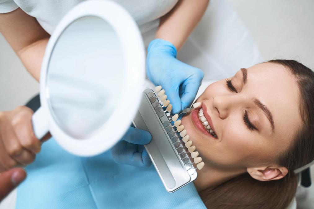 Dental professional matching tooth shade for a smiling patient using a shade guide and magnifying mirror.
