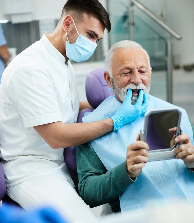 an elderly man is looking at his teeth in a mirror at the dentist 's office .