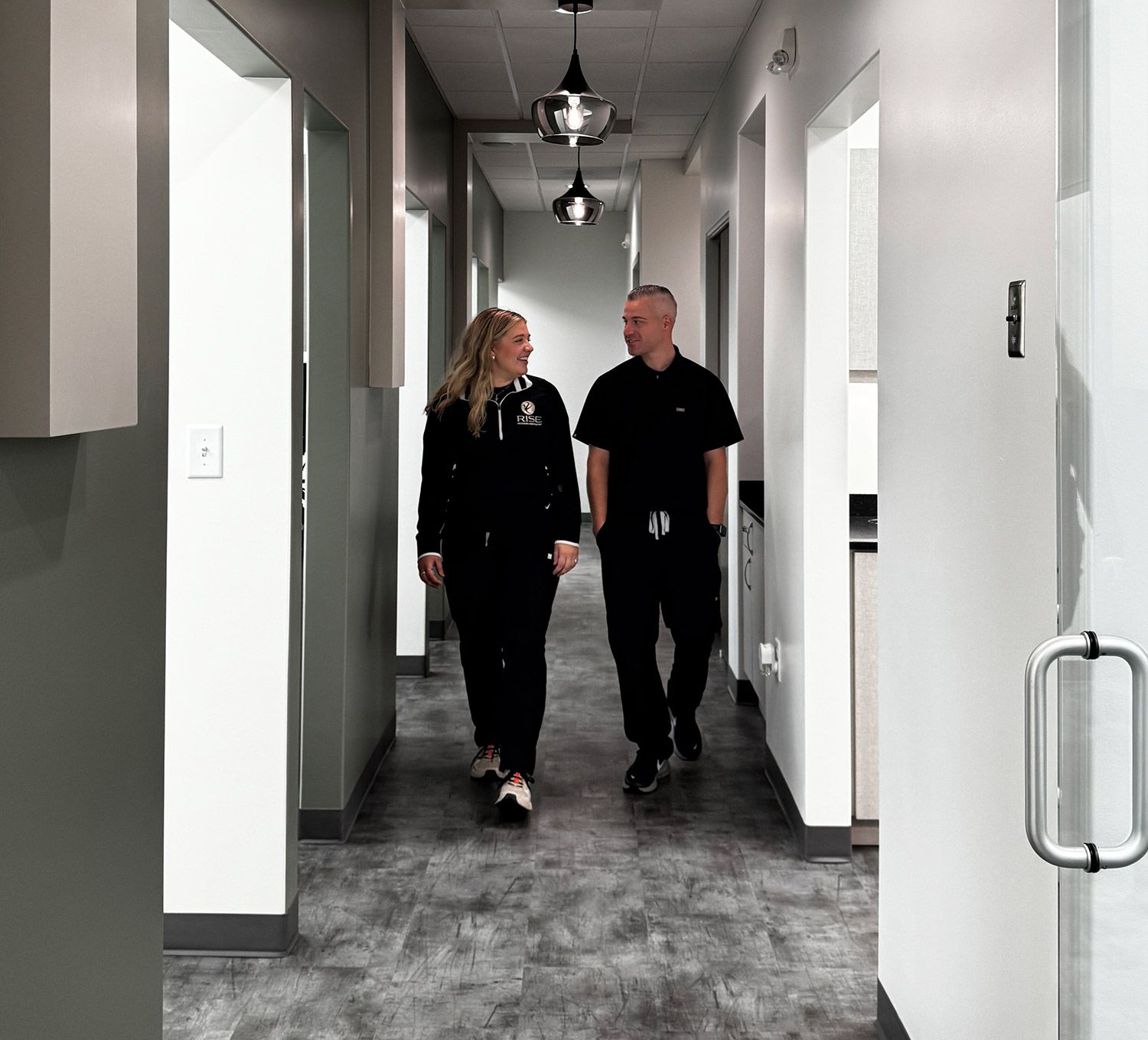 A man and woman in black scrubs smile while walking together in a modern hallway.