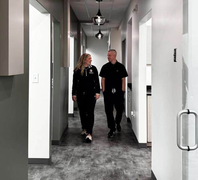 A man and a woman in black scrubs walk down a modern hallway, smiling at each other.