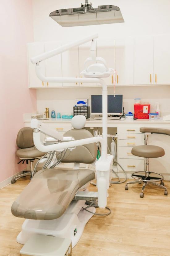 A modern dental operatory featuring a taupe dental chair, white cabinets with gold hardware, a light wooden floor, and a light pink wall.