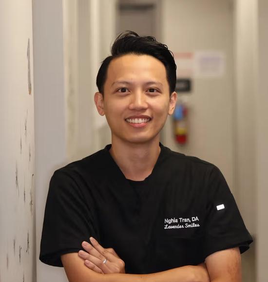 a man in a black scrub top is leaning against a wall with his arms crossed and smiling .