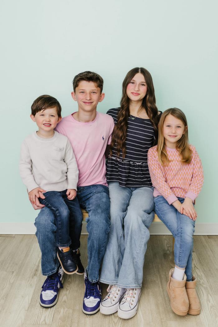 Four smiling children, two boys and two girls, sit on a bench against a light blue wall.