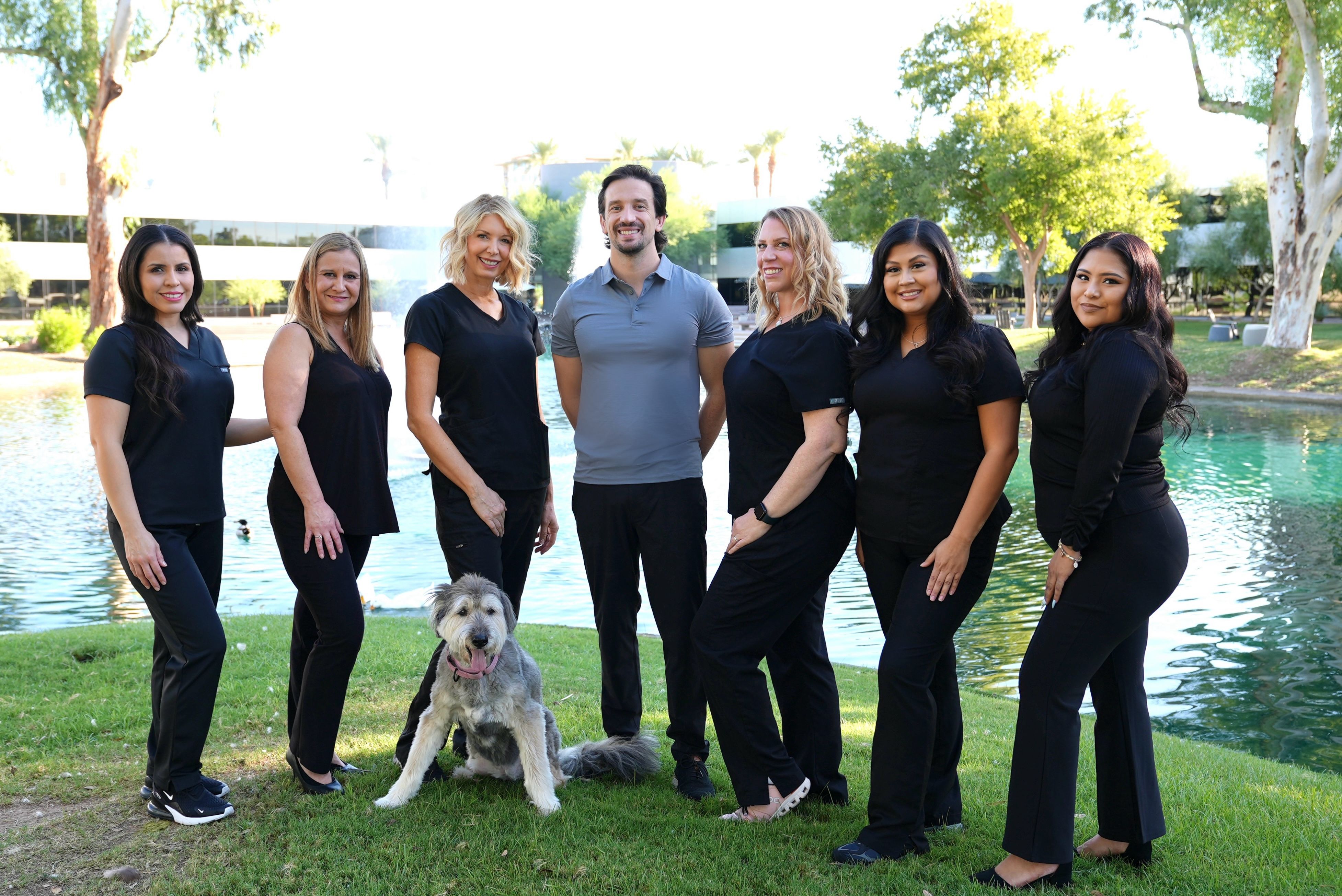 Seven smiling medical professionals and a fluffy grey dog pose on green grass beside a pond.