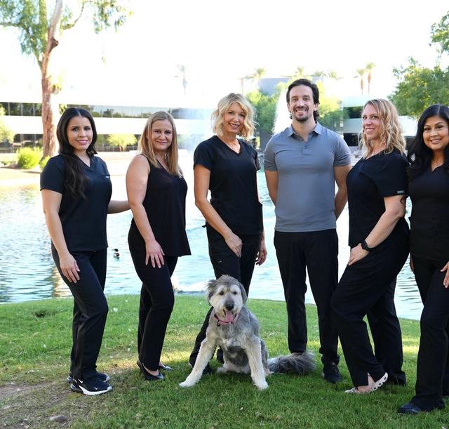 Seven smiling medical professionals and a fluffy grey dog pose on green grass beside a pond.