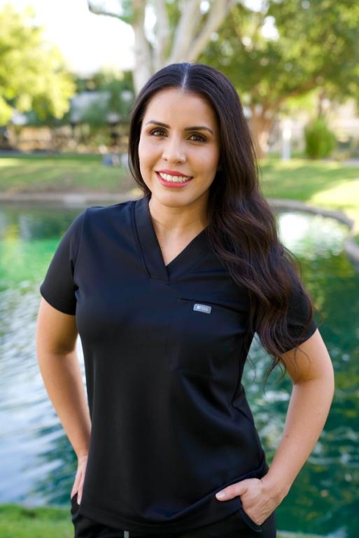 A smiling woman with long dark hair wears black FIGS scrubs, standing outdoors by a pond with greenery.