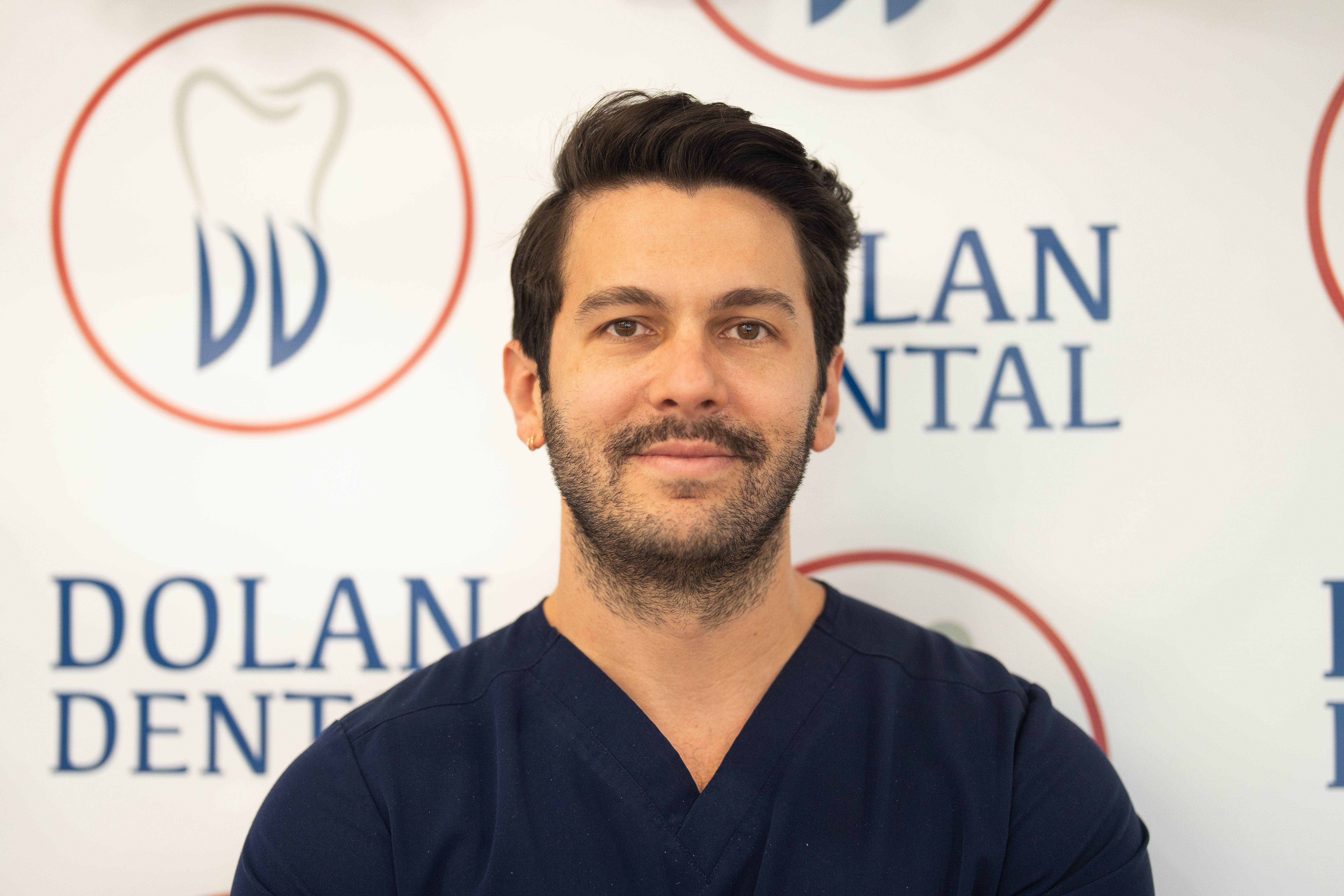 Smiling man in navy scrubs with a beard and mustache, in front of the Dolan Dental logo.