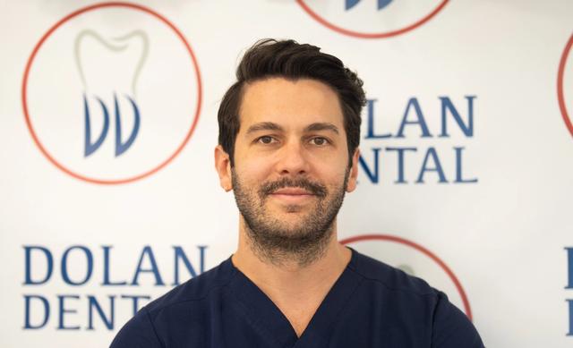 Smiling man in navy scrubs with a beard and mustache, in front of the Dolan Dental logo.