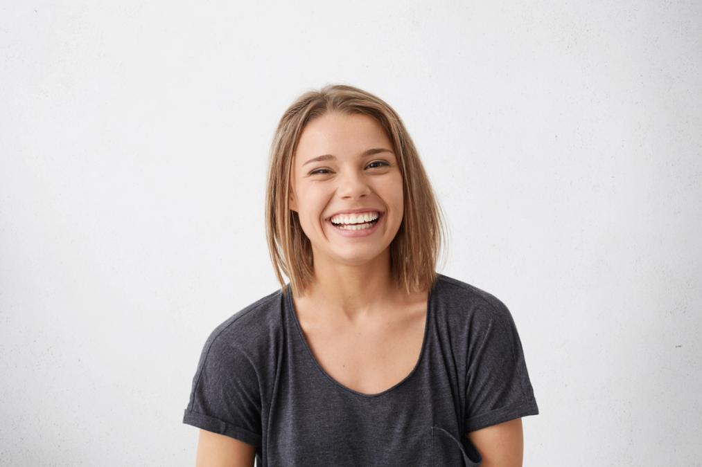 A young woman with short brown hair laughs openly, showing her teeth, against a white background.