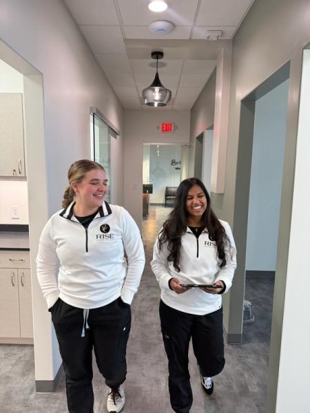 Two women in matching white "RISE" pullovers smile while walking down a hallway.