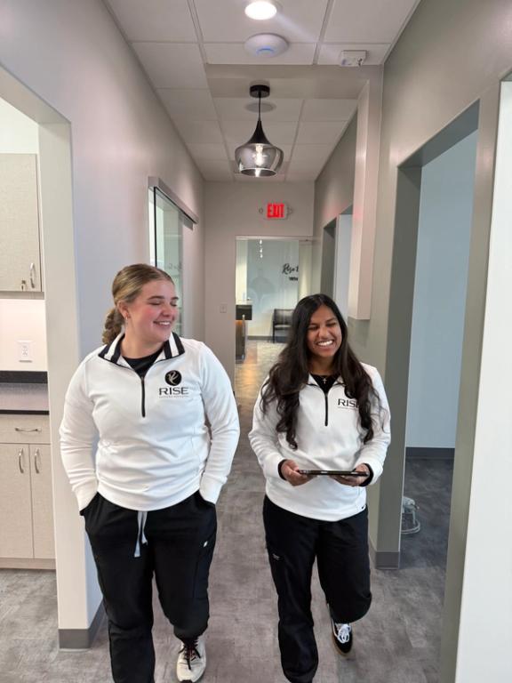Two smiling women in white tops and black pants walk down a hallway, one holding a tablet.