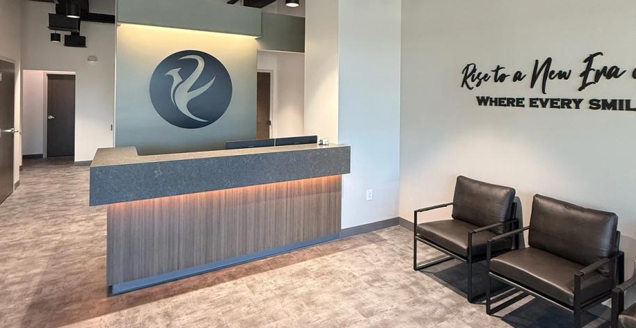 A modern reception area with a backlit front desk, a circular logo on the wall, and two dark waiting chairs.