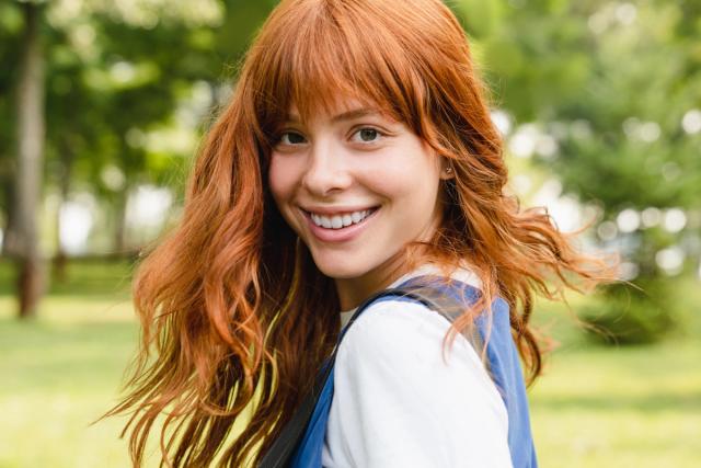 a woman with red hair and a backpack is smiling in a park .