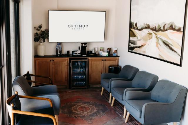 A modern dental office waiting room with gray armchairs, a screen displaying "Optimum Dental," and a beverage station.