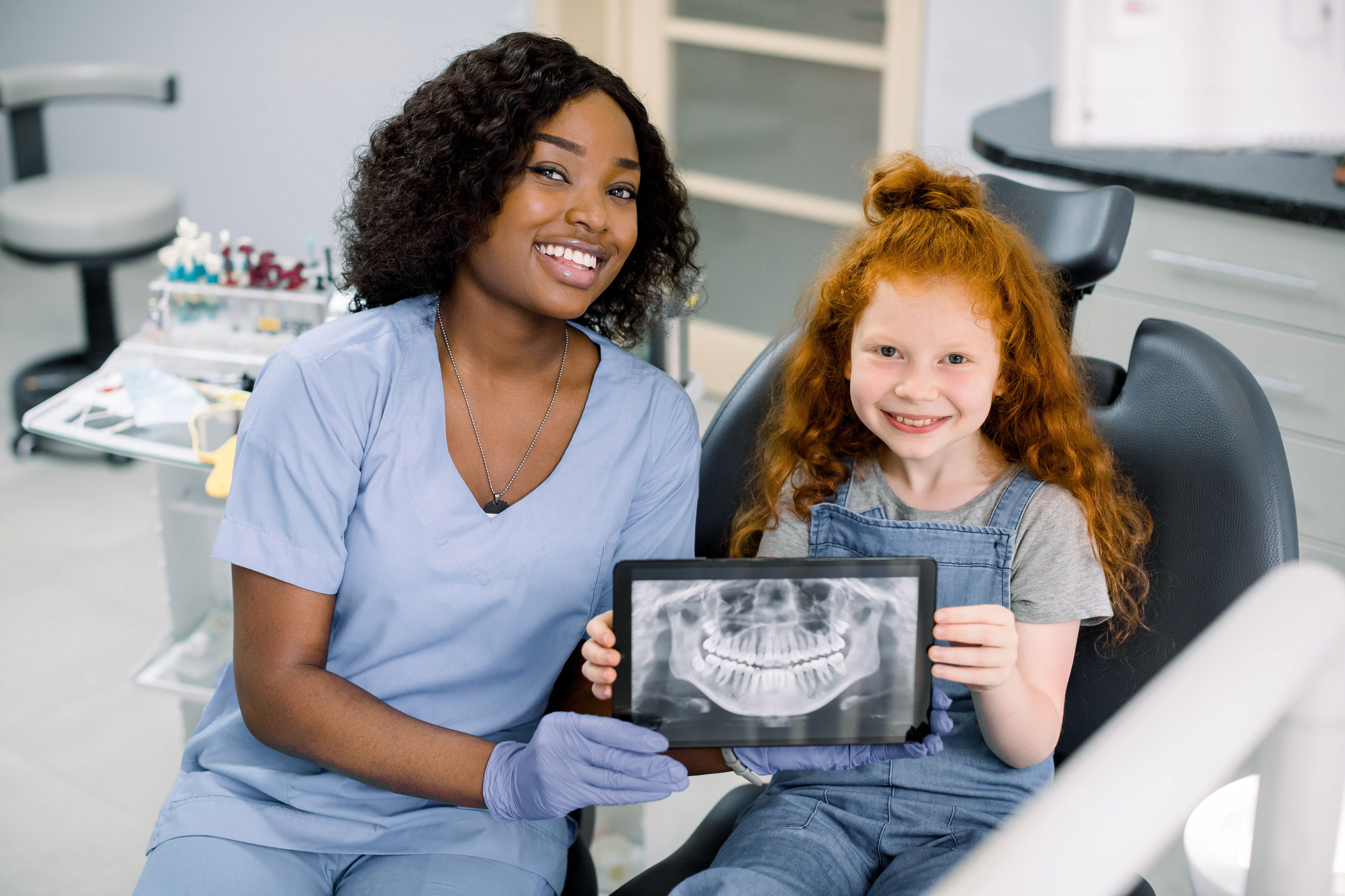 A Black dental professional and a red-headed girl smile, holding a tablet displaying a dental X-ray.