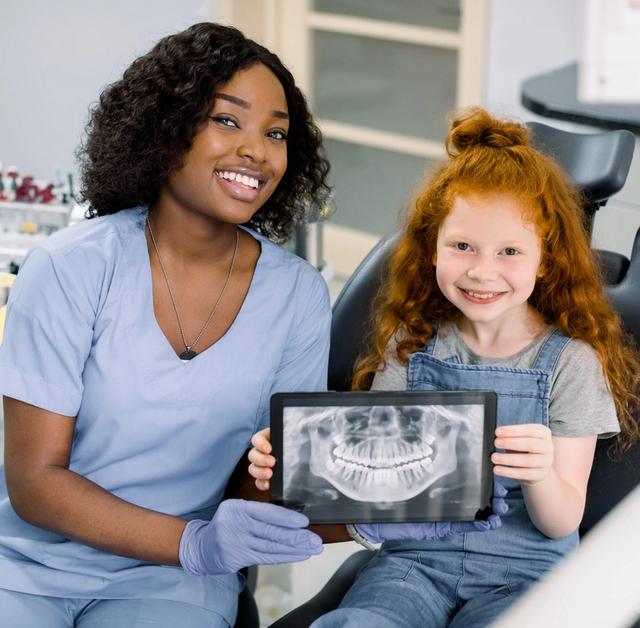 A Black dental professional and a red-headed girl smile, holding a tablet displaying a dental X-ray.