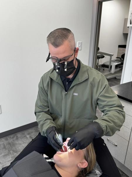A masked dentist with loupes performs a procedure on a patient in a dental chair.