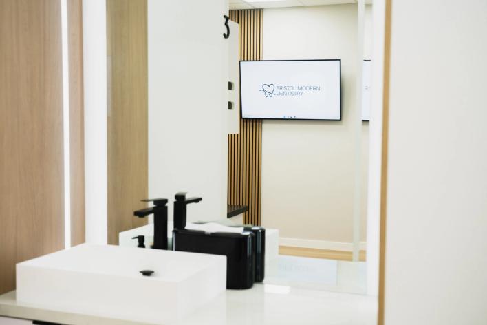 A modern white sink and black faucets, with a mirror reflecting a TV screen displaying the Bristol Modern Dentistry logo.