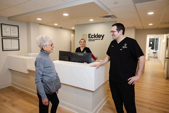 A smiling dentist greets an older female patient in a modern dental office lobby, with a receptionist smiling behind the desk.