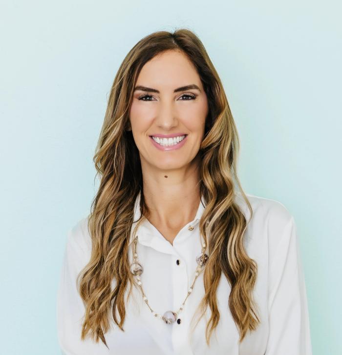 A smiling woman with long wavy hair, wearing a white shirt and necklace, against a light blue background.