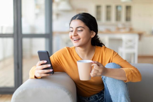 a woman is sitting on a couch holding a cup of coffee and looking at her phone .