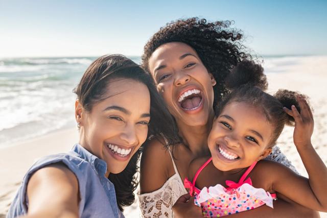 three women and a little girl are taking a selfie on the beach .