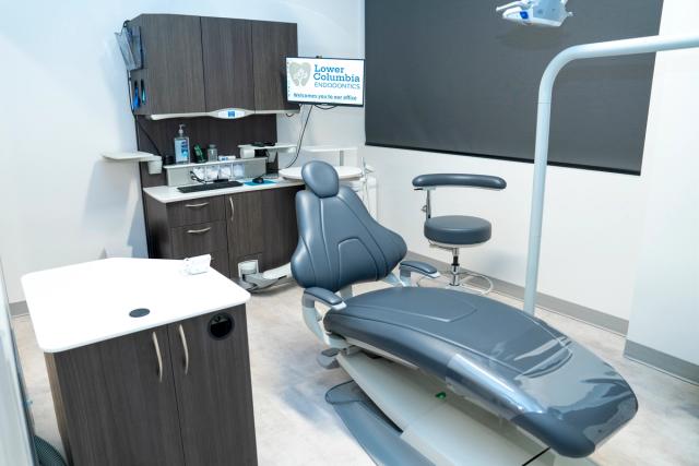 A modern dental examination room with a gray dental chair, dark wood cabinets, and a monitor displaying "Lower Columbia Endodontics."
