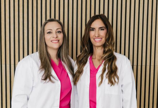 Two smiling women in white lab coats and hot pink scrubs against a wooden slat wall.