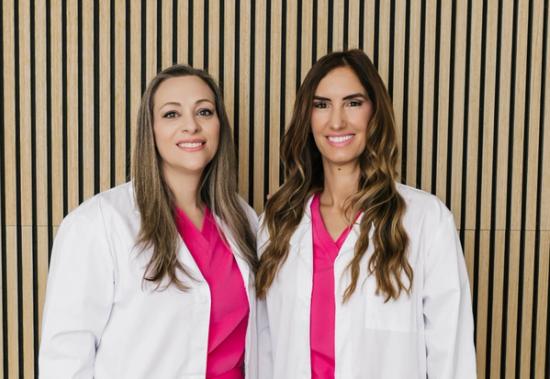 Two smiling women in white lab coats and hot pink scrubs against a wooden slat wall.