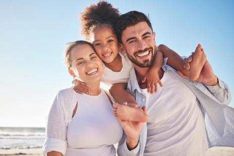 A family of three, with a daughter on her father's shoulders, smiling on a beach.